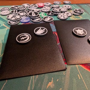 May include: A collection of black and white game tokens with various symbols, scattered on a patterned surface. Two black rectangular cards have tokens placed on them. The tokens are round with white outlines and symbols.