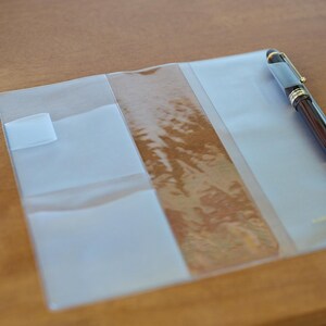 May include: A clear, rectangular plastic notebook cover featuring multiple interior pockets and flaps for organisation. A dark brown fountain pen with gold-coloured trim is tucked into the right-hand side pocket of the transparent cover.