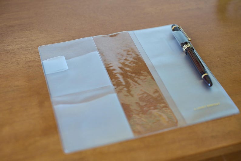 May include: A clear, rectangular plastic notebook cover featuring multiple interior pockets and flaps for organisation. A dark brown fountain pen with gold-coloured trim is tucked into the right-hand side pocket of the transparent cover.