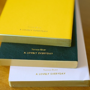 May include: A stack of three rectangular notebooks with gold-gilded edges. The top notebook is bright yellow, the middle is dark green, and the bottom is white. Each cover features the text Tomoe River and A LOVELY EVERYDAY in gold lettering.