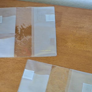 May include: Two clear, rectangular plastic covers for notebooks or planners. Each cover features multiple interior pockets and slots for organisation. The larger cover has the text A LOVELY EVERYDAY printed in small gold letters on the front panel.