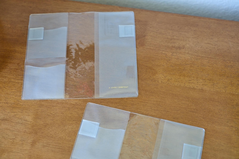 May include: Two clear, rectangular plastic covers for notebooks or planners. Each cover features multiple interior pockets and slots for organisation. The larger cover has the text A LOVELY EVERYDAY printed in small gold letters on the front panel.