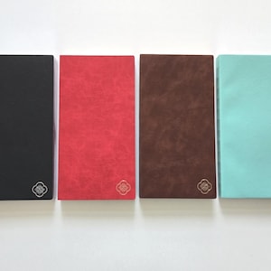 May include: Four rectangular notebooks in black, red, brown, and teal. Each notebook has a textured cover and a small, silver-colored floral emblem in the lower right corner. The notebooks are arranged side by side on a white surface.