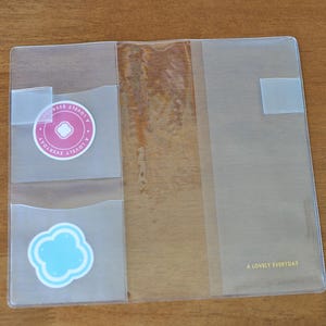 May include: A clear plastic notebook cover featuring two interior pockets on the left side. The top pocket contains a round pink sticker with the text A LOVELY EVERYDAY, and the bottom pocket holds a light blue cloud-shaped sticker.
