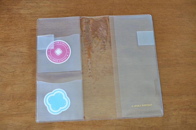 May include: A clear plastic notebook cover featuring two interior pockets on the left side. The top pocket contains a round pink sticker with the text A LOVELY EVERYDAY, and the bottom pocket holds a light blue cloud-shaped sticker.