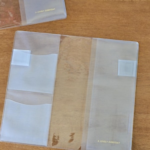 Könnte beinhalten: Eine durchsichtige Notizbuchhülle aus Kunststoff mit zwei Innentaschen auf der linken Seite und einem durchgehenden Fach auf der rechten Seite. Der Text A LOVELY EVERYDAY ist in kleinen goldenen Buchstaben unten rechts aufgedruckt.
