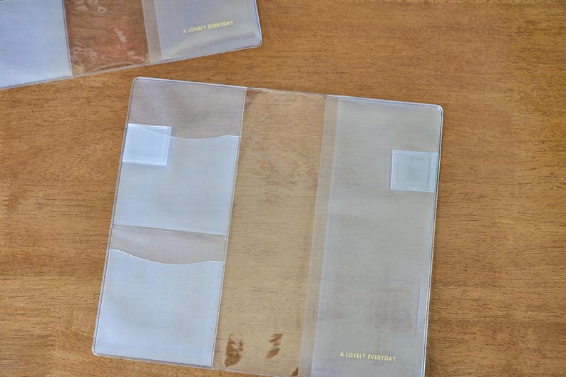 May include: A clear plastic notebook cover featuring two interior pockets on the left side and a full-length sleeve on the right. The text A LOVELY EVERYDAY is printed in small gold letters on the bottom right corner of the transparent cover.