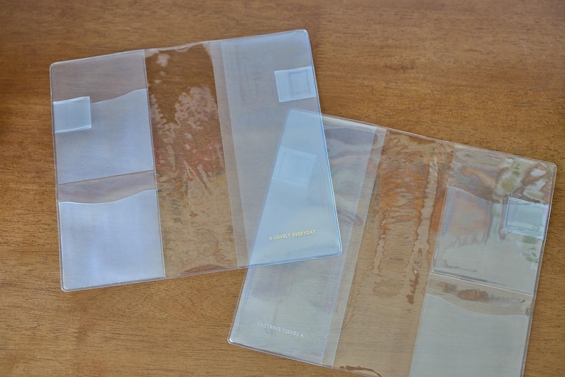 May include: Two clear plastic covers for notebooks or planners, each featuring multiple interior pockets and slots for organisation. The transparent material allows for visibility of contents, and the design includes small rectangular tabs.