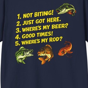 May include: A navy blue t-shirt featuring yellow text that reads: 1. NOT BITING! 2. JUST GOT HERE. 3. WHERE'S MY BEER? 4. GOOD TIMES! 5. WHERE'S MY ROD? The design includes three colorful illustrations of fish positioned around the printed text.