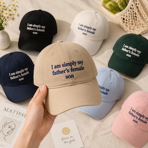 May include: A collection of baseball caps in various colors, including black, white, navy, tan, green, light blue, and pink. Each cap features the embroidered text "I am simply my father's female son."