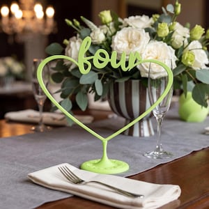 May include: A lime green heart-shaped table number holder with the word "four" in cursive script. The holder is on a table set for a formal event, with a white napkin and fork in the foreground and a floral arrangement in the background.