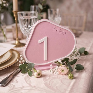 May include: A pink table number with a white border and the number 1. The word "TABLE" is embossed at the top. The table number is decorated with flowers and greenery, set on a table with a floral tablecloth.