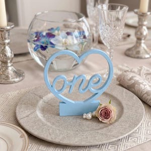 May include: A light blue wooden table decoration in the shape of a heart with the word "one" in cursive. The decoration is on a gray plate with a pink rose and pearls. A glass bowl with flowers is in the background.