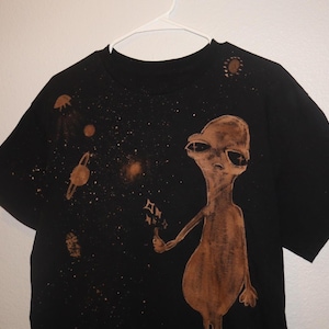 Puede incluir: Camiseta negra con un extraterrestre marrón sosteniendo una estrella. El diseño incluye planetas y estrellas, creando un tema espacial. La camiseta está colgada de una percha blanca.