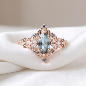 May include: Rose gold ring with a large, light blue marquise-cut gemstone. The ring is decorated with smaller white and blue gemstones in a floral pattern. The ring is set on a white satin surface.