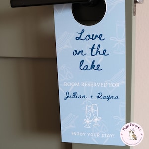 May include: A light blue door hanger with the text "Love on the lake" in cursive. The words "Room Reserved For" and the names "Jillian + Rayna" are also visible. The bottom of the hanger reads "Enjoy Your Stay!"
