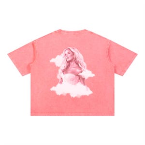 May include: Coral-coloured t-shirt with a portrait of a woman with long blonde hair, wearing a white corset, and surrounded by clouds. The image is printed on the back. The shirt has short sleeves and a crew neck.