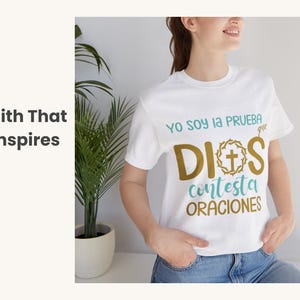 May include: White t-shirt with the Spanish text "Yo soy la prueba que Dios contesta oraciones" in teal and gold lettering. The design includes a cross and a wreath. The shirt is a casual wear item.