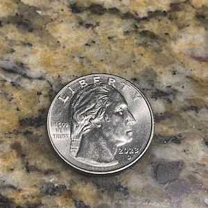 Może przedstawiać: Amerykańska moneta ćwierćdolarowa, z datą 2023 roku, z profilem George'a Washingtona skierowanym w prawo. Moneta zawiera napisy "LIBERTY" i "IN GOD WE TRUST". Moneta jest srebrna.