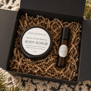 May include: A black gift box containing a jar of "Nordic Botanicals Body Scrub" (4 oz) and a roller bottle of "Lavender Chamomile Roller" (10 ml). The products sit on brown paper filler. The box is open, revealing the contents.