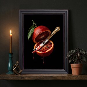 May include: A framed wall art print featuring a halved blood orange with a green leaf, pierced by an ornate gold-toned knife. Dark red juice drips from the fruit onto the blade, where several small black flies are perched against a black background.