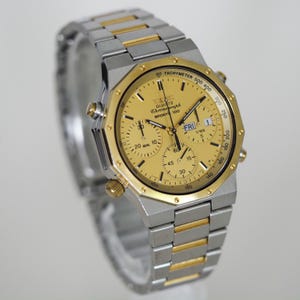 May include: A two-tone Seiko Quartz Chronograph Sports 100 watch with a gold-colored dial and a silver and gold-tone stainless steel link bracelet. The watch features a tachymeter bezel, three sub-dials, and a day-date window at the three o'clock position.