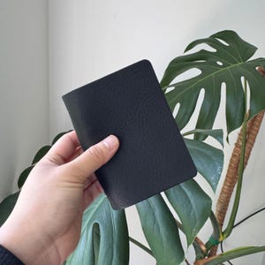 Op de afbeelding: Een zwart, getextureerd leren notitieboekje in een hand. Het notitieboekje is rechthoekig met afgeronde hoeken. De achtergrond bevat grote groene bladeren van een kamerplant.