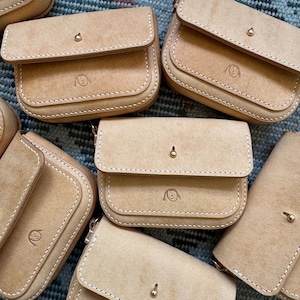 May include: Several small, light tan leather pouches with rounded edges and a flap closure. Each pouch has a small, gold-coloured clasp and a stitched design. A simple illustration is embossed on the front of each pouch.