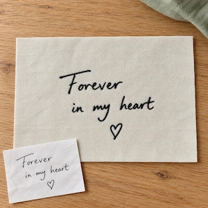 May include: A rectangular piece of off-white fabric featuring black embroidered text that reads "Forever in my heart" with a small heart symbol below. A small white paper note with the same handwritten text is placed in the corner.