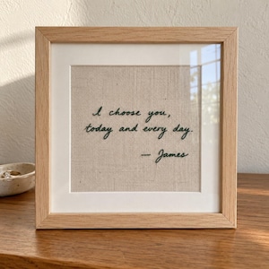 May include: A square wooden frame containing a piece of light beige fabric with dark green embroidered text that reads, I choose you, today and every day. -- James. The embroidery is centered on the fabric, which is set behind clear glass.