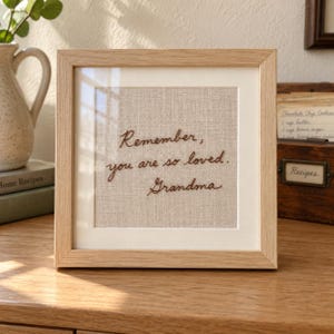 Puede incluir: Un marco de madera cuadrado que contiene una tela de color beige claro con texto bordado en cursiva de color marrón oscuro que dice: Remember, you are so loved. Grandma. El bordado está centrado sobre la tela, detrás de un paspartú blanco.