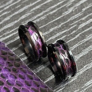 Peut inclure: Deux tunnels d'oreille en métal noir avec un motif incrusté ressemblant à une peau de serpent violette et noire. Le bijou est placé à côté d'un morceau de matière texturée en peau de serpent violette. Le texte Serpen Stitch est visible en haut.