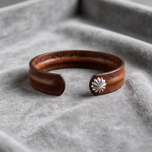 May include: A brown leather cuff bracelet featuring a smooth, rounded texture and a single silver-toned metal concho stud accent at one end. The bracelet has an open design with a curved, flexible band that displays natural leather grain.