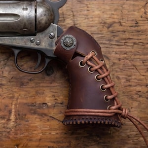 May include: A metallic revolver featuring a dark brown leather grip cover. The leather wrap is secured with brass-toned eyelets and a matching leather lace, finished with a decorative silver-toned metal concho button near the frame.