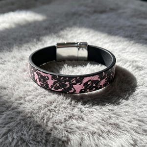 May include: A bracelet featuring a black and pink snakeskin-style pattern on the exterior band. The interior of the band is solid black, and it is secured with a rectangular, polished silver-toned metal magnetic clasp closure.