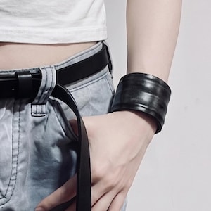 Puede incluir: Una pulsera de puño ancha y lisa de color negro con una apariencia ligeramente curvada de varias bandas. La pulsera presenta un acabado oscuro y sólido que se envuelve alrededor de la muñeca, ofreciendo una pieza accesoria audaz.