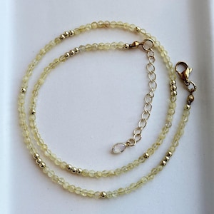 May include: A delicate necklace featuring small, faceted pale yellow gemstone beads interspersed with polished gold-tone metal beads. The necklace includes a gold-tone lobster claw clasp and an adjustable chain extender finished with a small, iridescent oval charm.