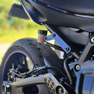 May include: A motorcycle rear section featuring a black seat, a blue metal passenger footpeg bracket with the Yamaha logo, and a small translucent brake fluid reservoir with orange fluid connected by a black hose, positioned above the rear tire.