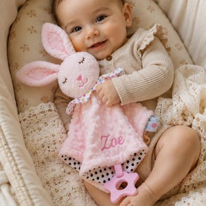 May include: A soft pink baby security blanket featuring a plush rabbit head with embroidered closed eyes and a nose. The blanket has a dimpled texture, a floral collar, a black and white polka dot underside, and a pink star-shaped teether.