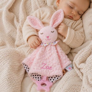 May include: A pink plush security blanket featuring a rabbit head with embroidered closed eyes and a nose. The fabric has a textured dot pattern, a colorful polka dot collar, and black and white polka dot backing. The name Zoe is embroidered in pink.