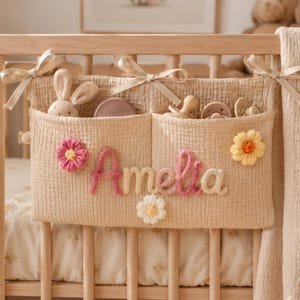 May include: A beige muslin crib organizer with two pockets, tied to a wooden crib with fabric ribbons. The name Amelia is embroidered in pink and cream yarn, decorated with three crocheted flowers in pink, yellow, and white. It holds a small plush rabbit and several pacifiers.