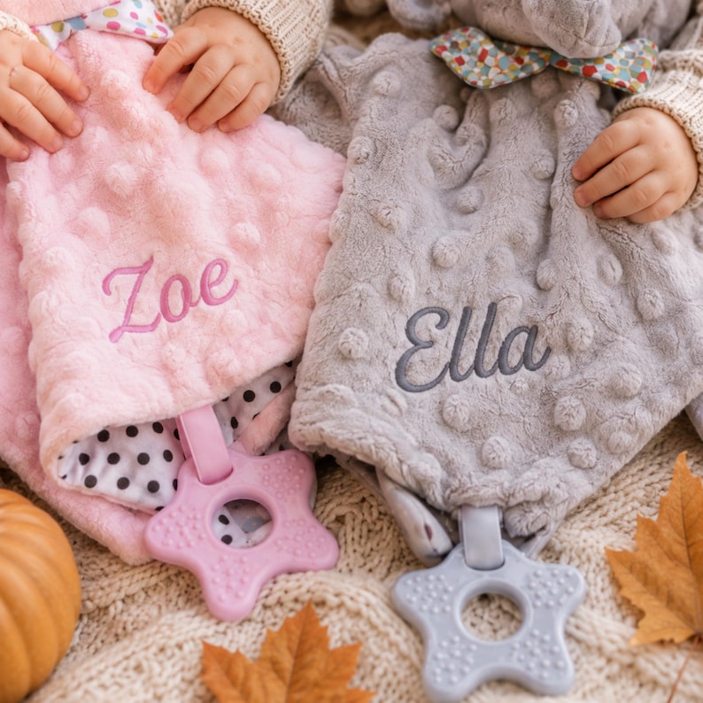 May include: Two soft plush security blankets with dimpled textures, one in light pink embroidered with the name Zoe and one in light gray embroidered with the name Ella. Each blanket features a colorful patterned bow and a star-shaped plastic teether.