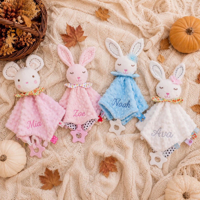 May include: Four plush security blankets featuring animal heads with embroidered faces. Each blanket has a soft dimpled texture, a small collar, and a star-shaped teether at the bottom. The names Mia, Zoe, Noah, and Ava are embroidered on the fabric.