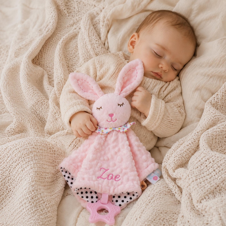 May include: A pink plush security blanket featuring a rabbit head with embroidered closed eyes and a nose. The fabric has a textured dot pattern, a colorful polka dot collar, and black and white polka dot backing. The name Zoe is embroidered in pink.