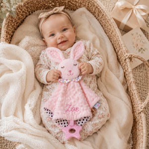 May include: A pink plush bunny security blanket featuring a textured dimpled fabric, a floral collar, and a star-shaped teether at the bottom. The name Zoe is embroidered in pink thread on the front. The blanket has a black and white polka dot lining.