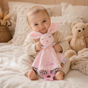 May include: A soft pink plush security blanket featuring a rabbit head with embroidered closed eyes and a nose. The blanket has a dimpled texture, a colorful patterned collar, and the name Zoe embroidered in pink. A star-shaped teether is attached.