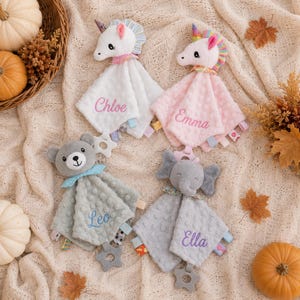 May include: Four plush security blankets featuring animal heads: two unicorns in white and pink, a gray bear, and a gray elephant. Each blanket has a dimpled texture, a star-shaped teether, and embroidered names: Chloe, Emma, Leo, and Ella.