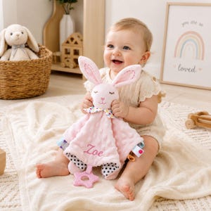 May include: A soft pink plush security blanket featuring a rabbit head with embroidered closed eyes and a nose. The fabric has a dimpled texture, a colorful polka dot neck ruffle, and the name Zoe embroidered in pink. A pink star-shaped teether is attached.
