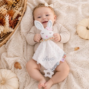 May include: A white plush security blanket featuring a stuffed bunny head with embroidered eyes, a pink nose, and a floral bow. The soft, dimpled fabric is embroidered with the name Ava in gray script and includes a white star-shaped teether.
