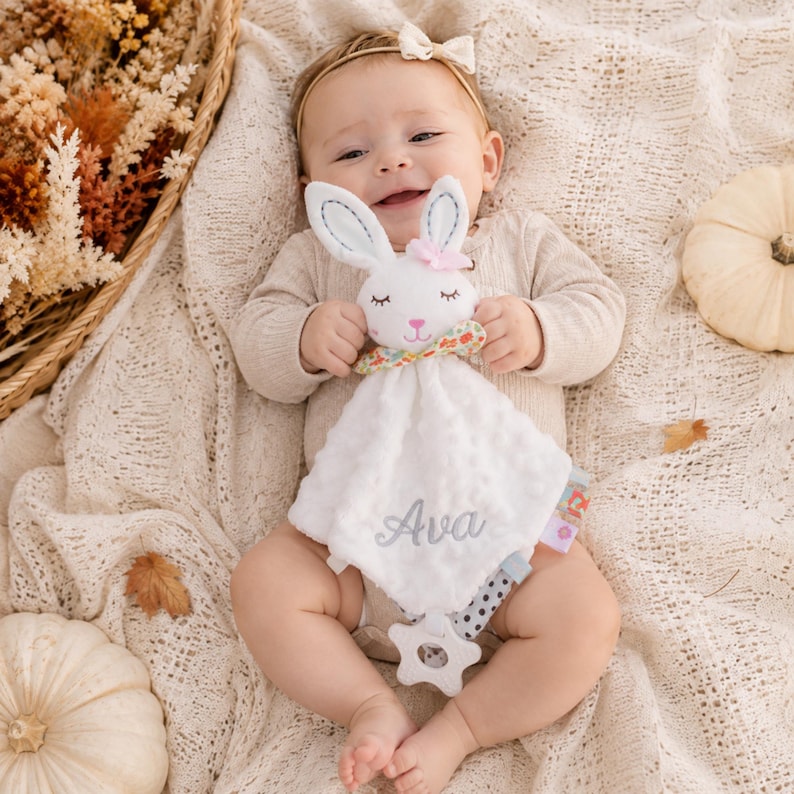May include: A white plush security blanket featuring a stuffed bunny head with embroidered eyes, a pink nose, and a floral bow. The soft, dimpled fabric is embroidered with the name Ava in gray script and includes a white star-shaped teether.